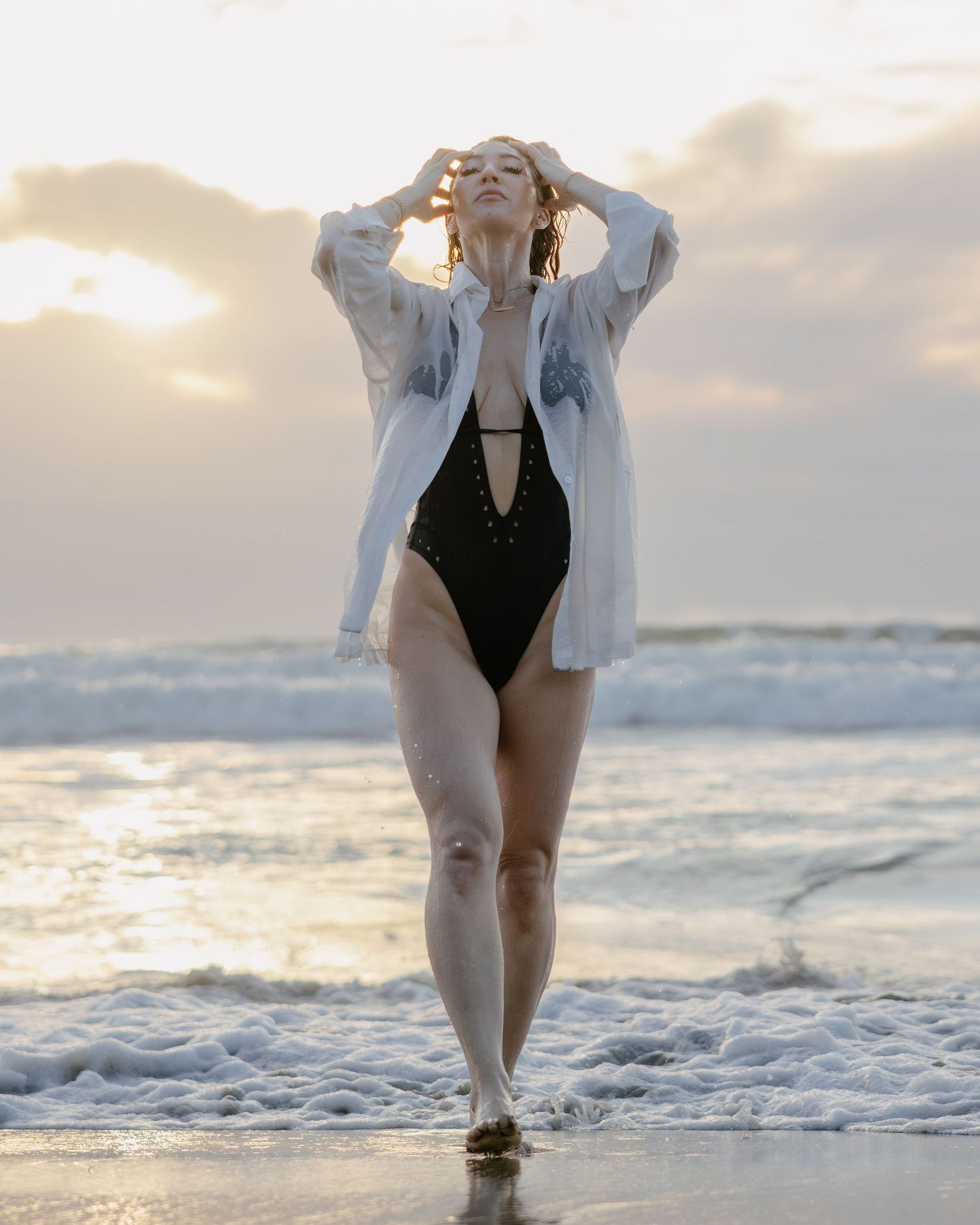 Figure in a white oversized shirt and black swimsuit poses on a beach at sunset with waves crashing in the background.