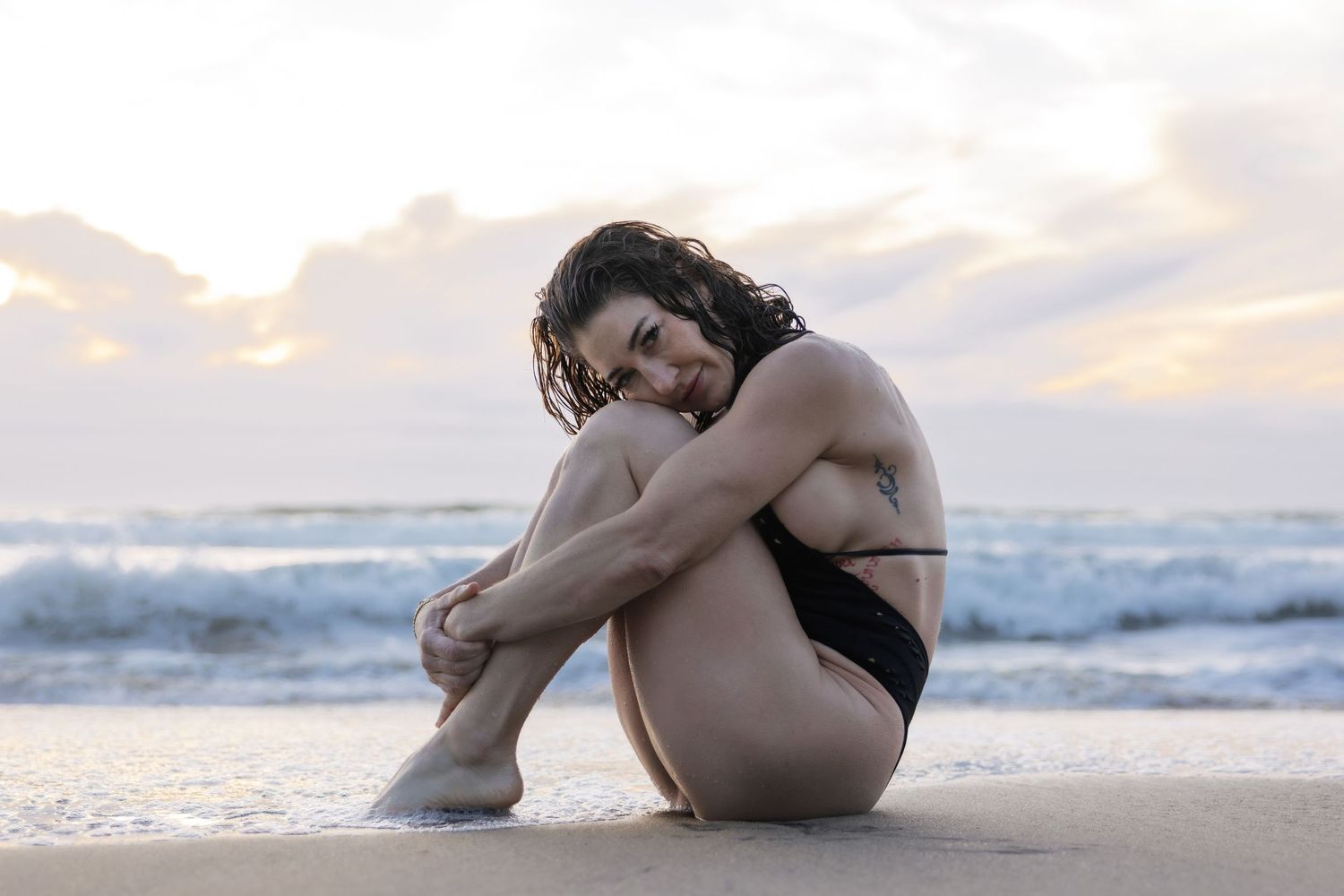 Someone in a black swimsuit sits thoughtfully on the beach at sunset as waves roll in behind them.