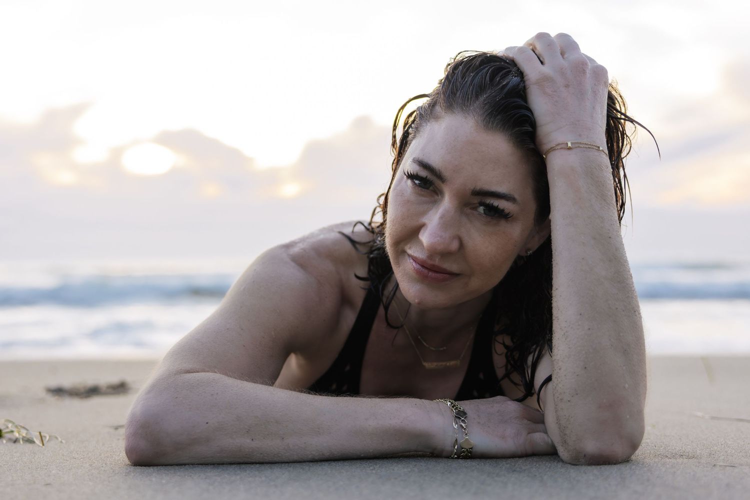 Someone with dark hair lies on a sandy beach at sunset smiling at the camera with an ocean view in the background.
