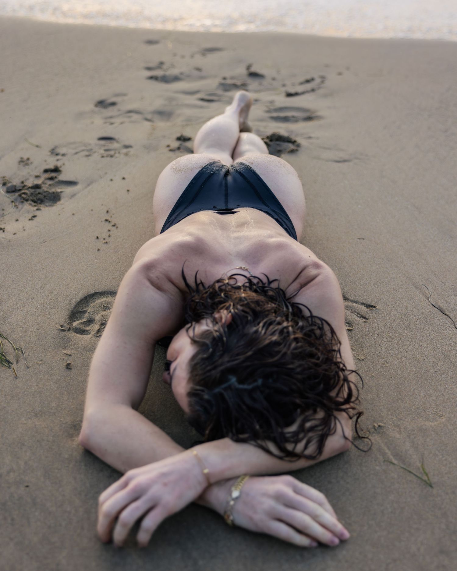 Person lying on sandy beach wearing dark swimsuit with curly hair resting on warm sand.