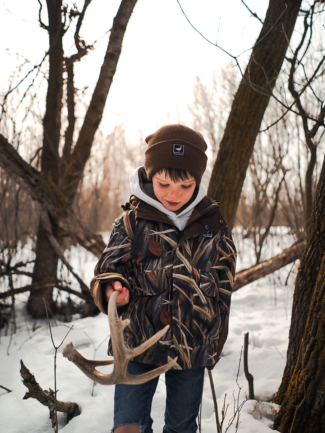 Young hunter in camouflage examining deer antlers in snowy woods.
