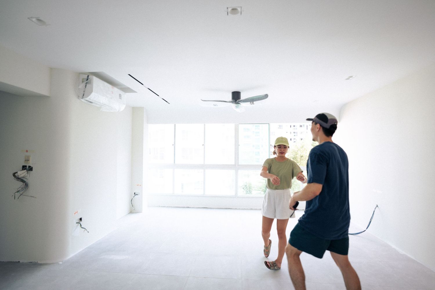 Two people stand in a bright, empty white room with a ceiling fan and large windows during daytime.