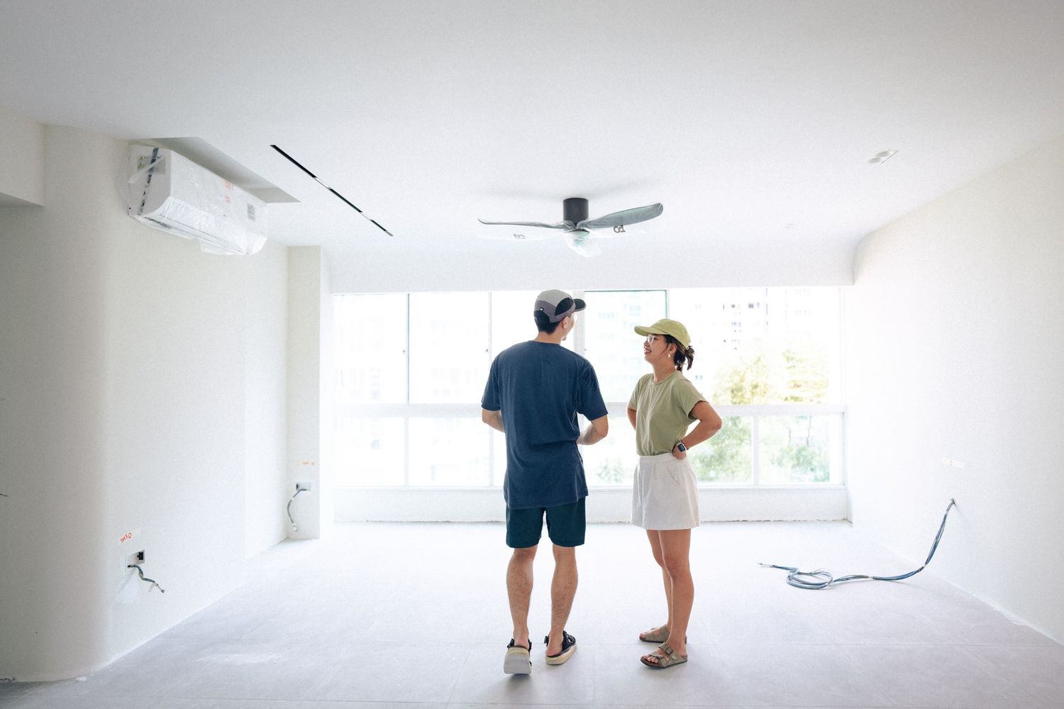 Two people viewing an empty white room with air conditioning unit and ceiling fan during property inspection.