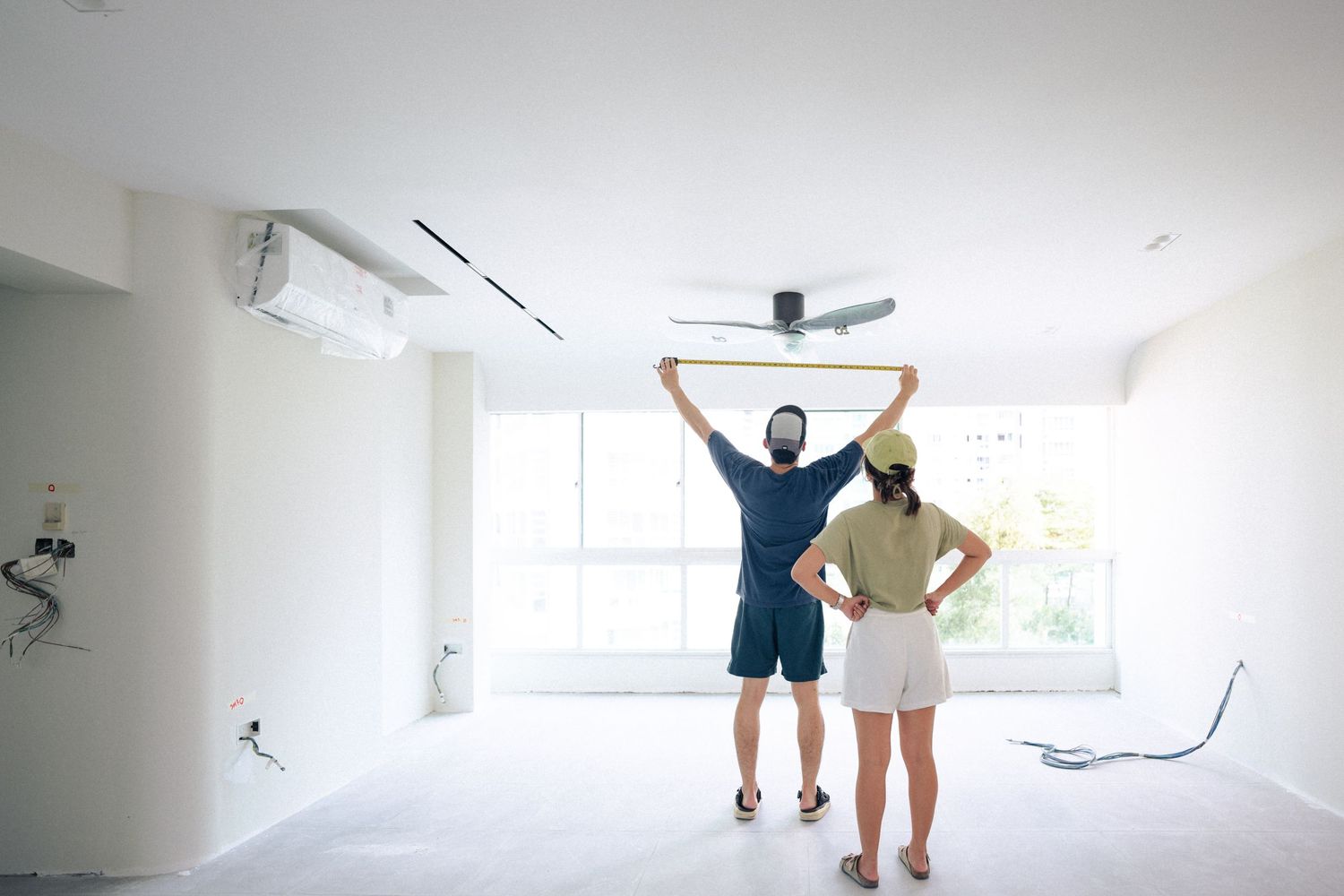 Two people standing in an empty white room with unfinished walls and ceiling, one person holding up construction materials.