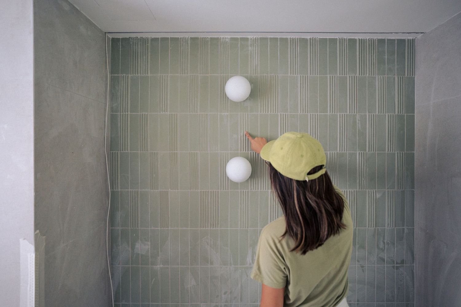 Person wearing a yellow cap looks up at two white spherical lights mounted on a gray tiled wall.