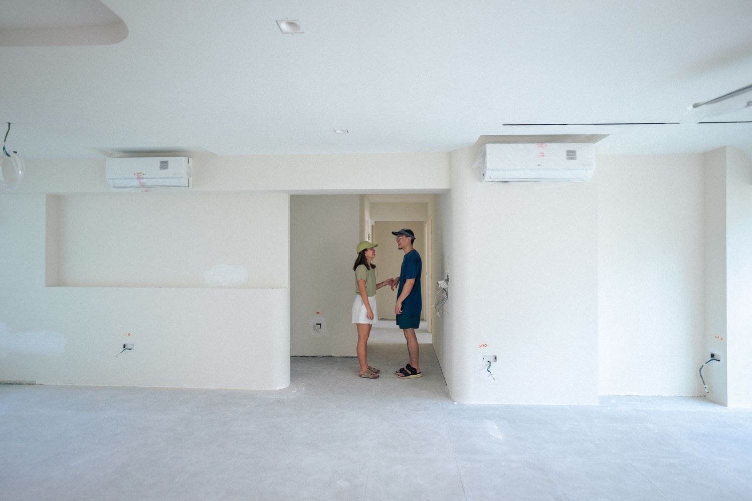 Two people inspecting a newly renovated empty white room with air conditioning units mounted on the walls.