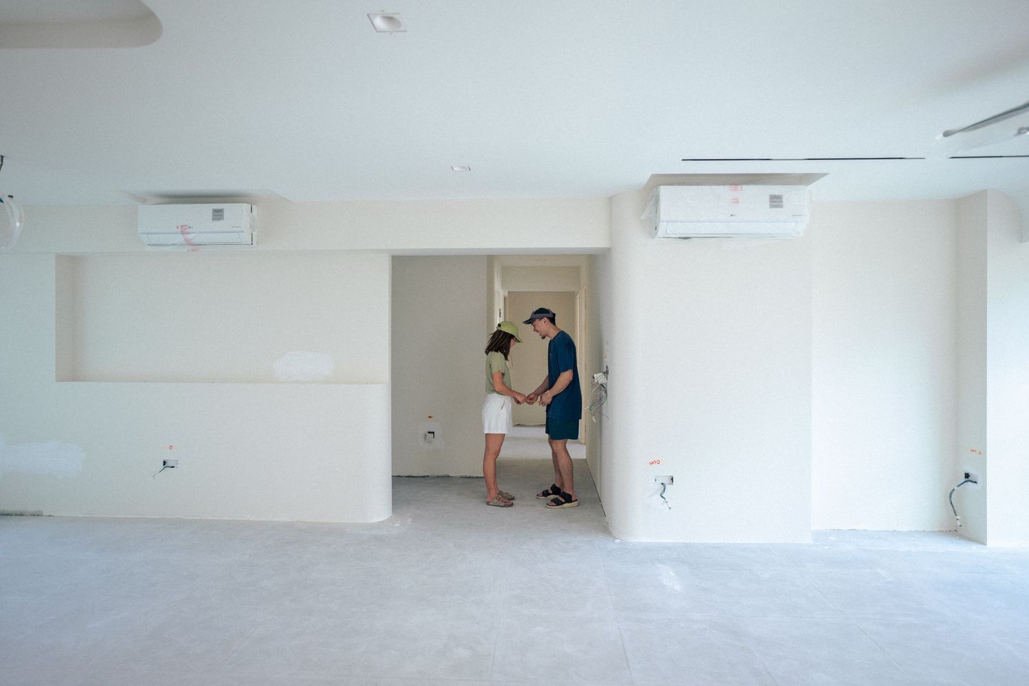 Two people stand together in a minimalist white empty room with air conditioning units mounted on the walls.