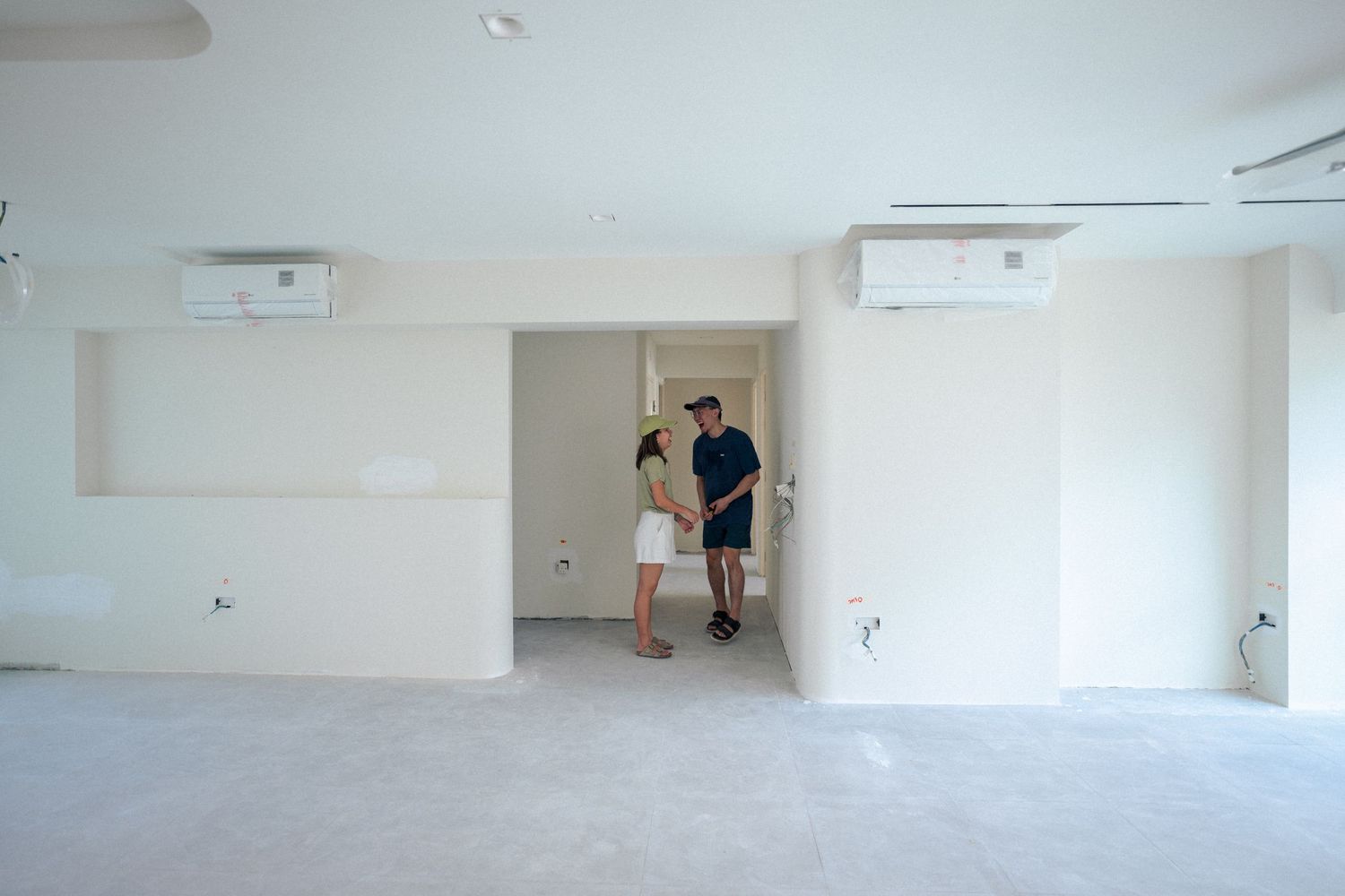 Two people stand in minimalist white empty room with air conditioning units visible on the wall.