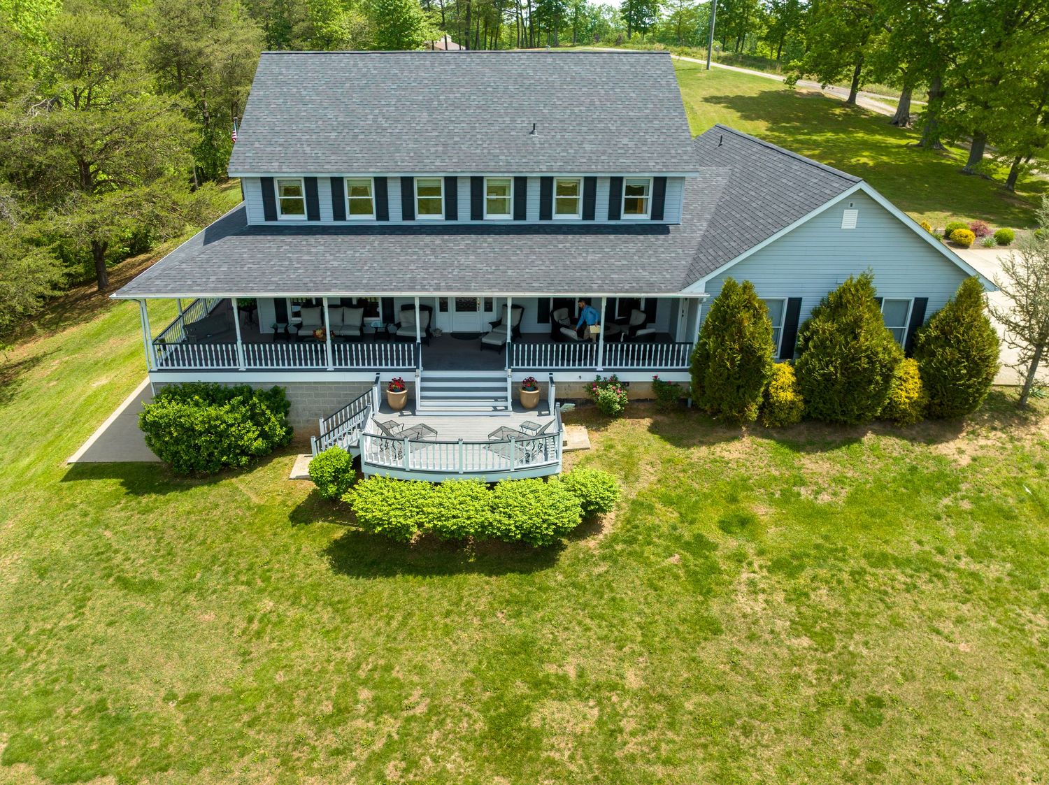 Aerial view of a spacious country home with white siding, green shutters, wraparound porch, and landscaped front yard.