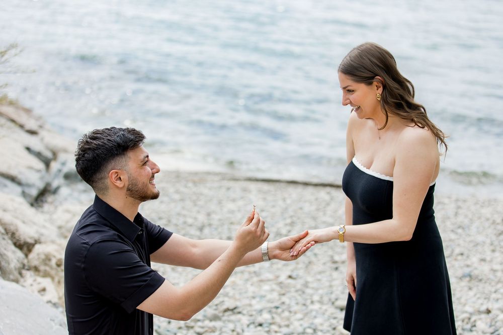 Man proposing to woman on pebble beach with water in background.