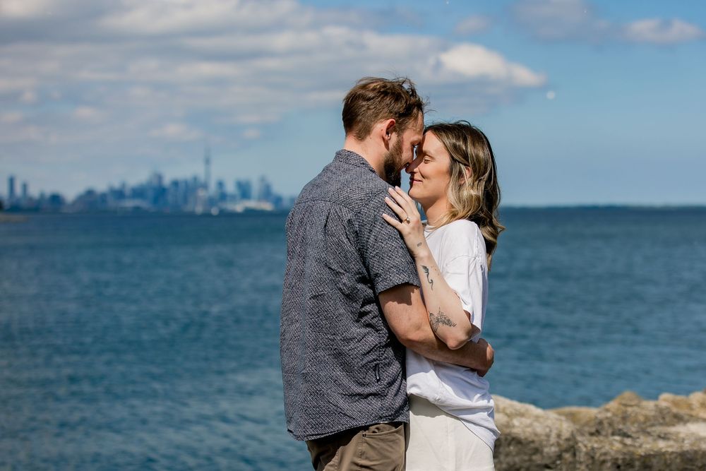 Romantic couple embraces by the ocean with city skyline visible in the background on a sunny day.