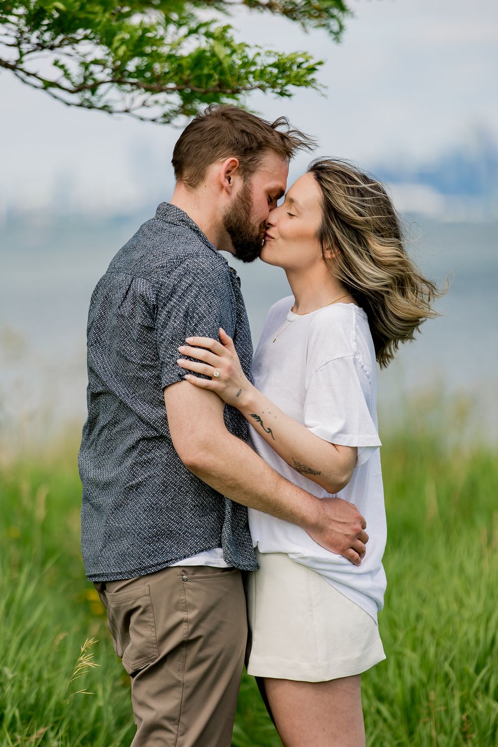 A romantic couple sharing an intimate kiss in a grassy meadow with cloudy skies in the background.