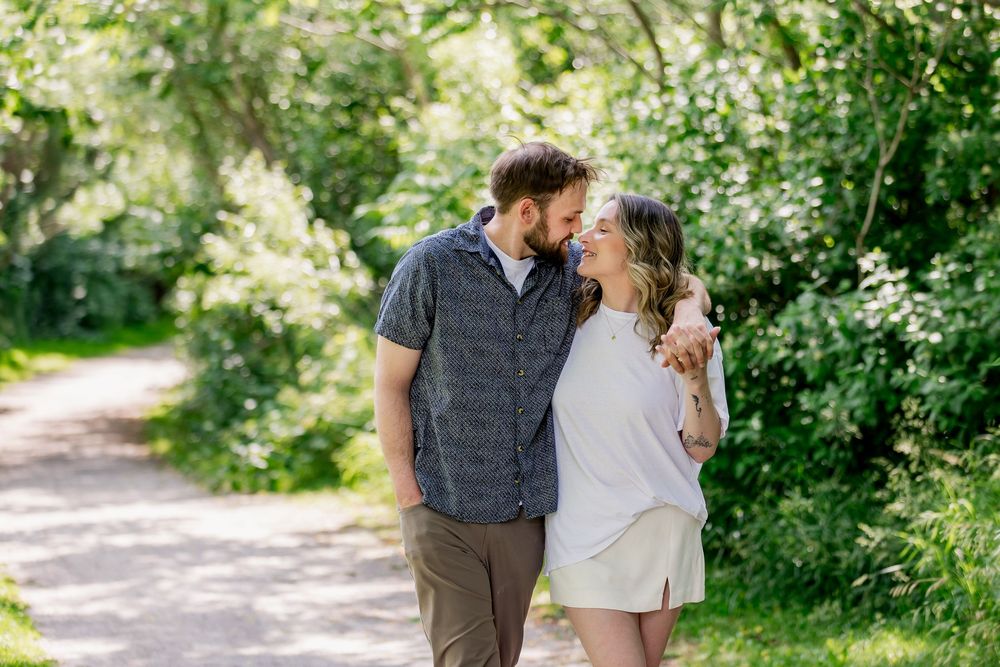 Couple sharing an intimate moment while walking on a sunlit gravel path bordered by lush summer greenery.