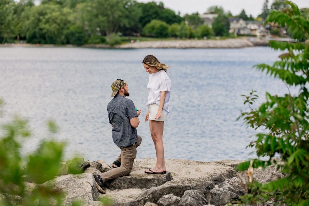 A romantic proposal moment unfolds on coastal rocks by a calm lake on a peaceful spring day.