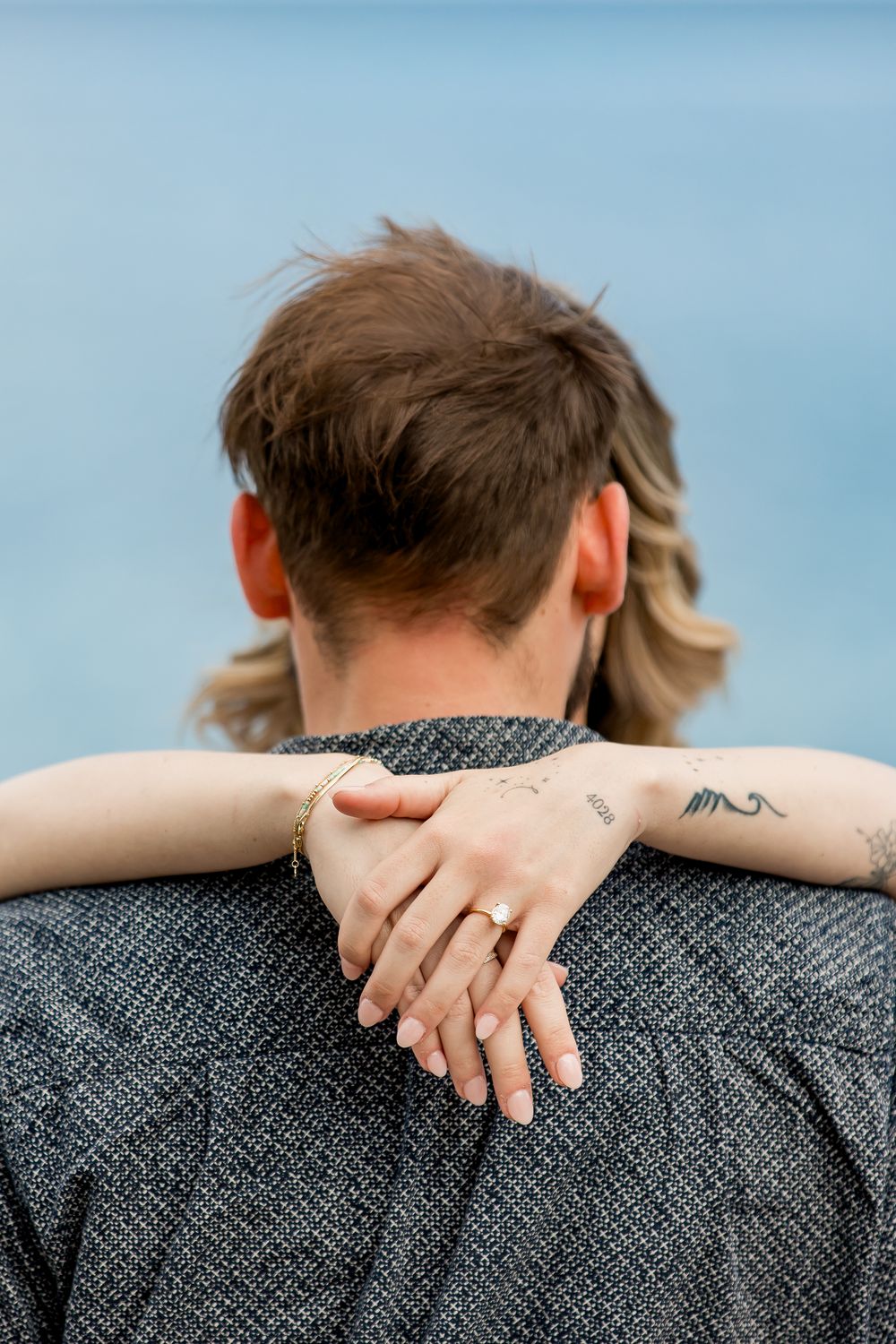 Close up view of hands with small tattoos embracing across shoulders wearing a gray textured sweater against blue sky.