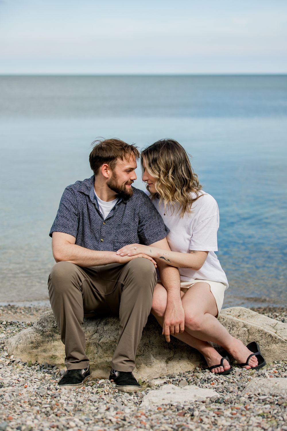 A couple sits together on a rocky beach shore sharing a tender moment by the calm blue water.