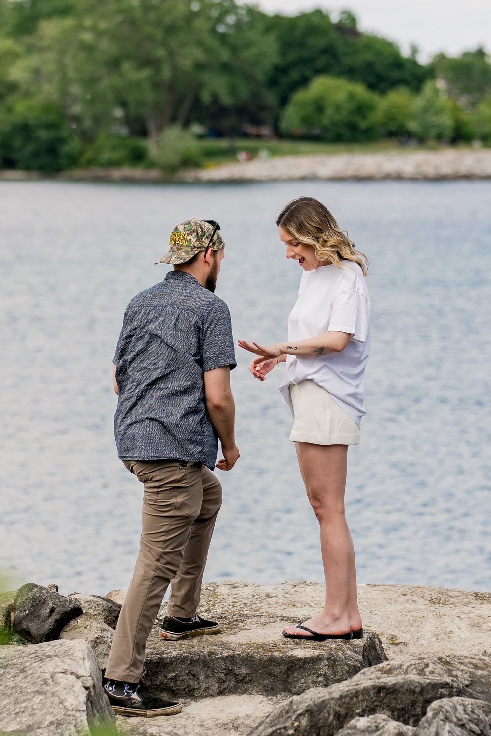 A couple stands on rocky shoreline by a lake during what appears to be a proposal moment.