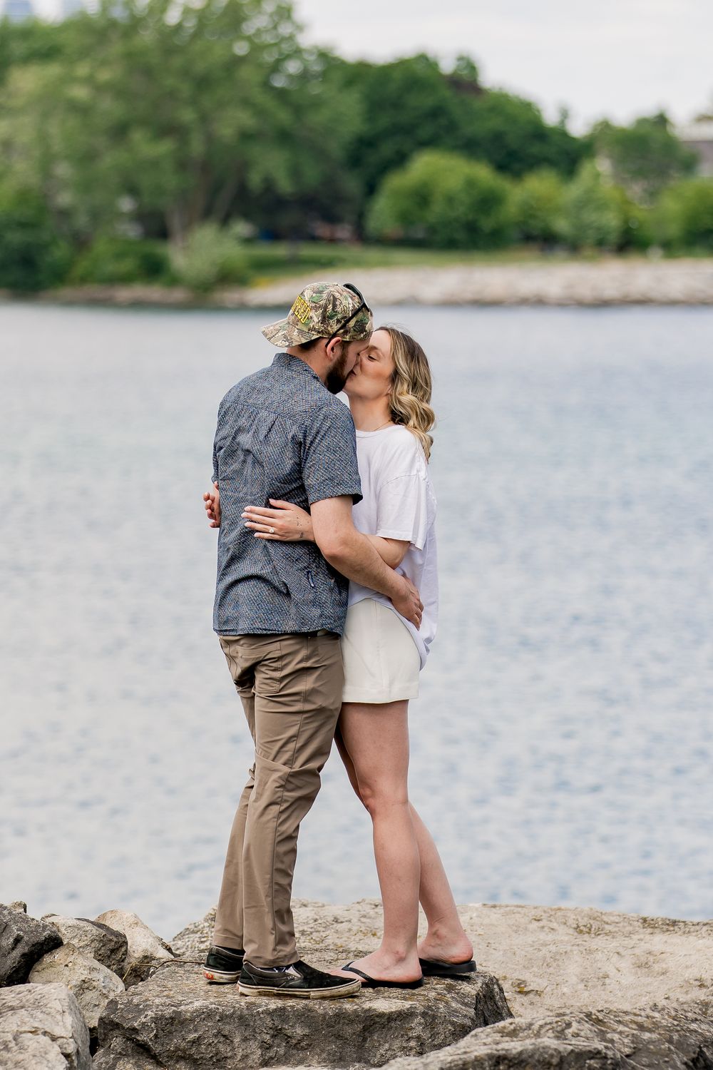 A couple shares a romantic moment while standing on rocks by a serene lake on a summer day.