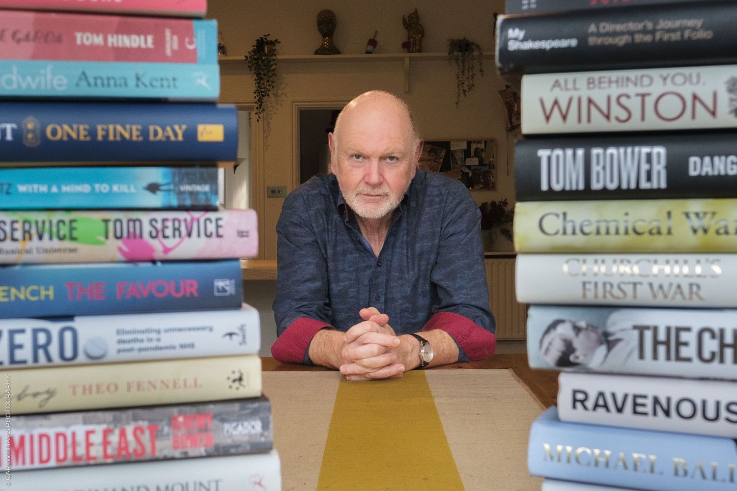 A person sits at a wooden table between two stacks of books in what appears to be a home library or study.