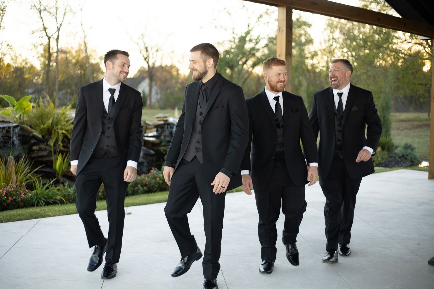 Groomsmen walk and chat while wearing formal black suits outdoors.