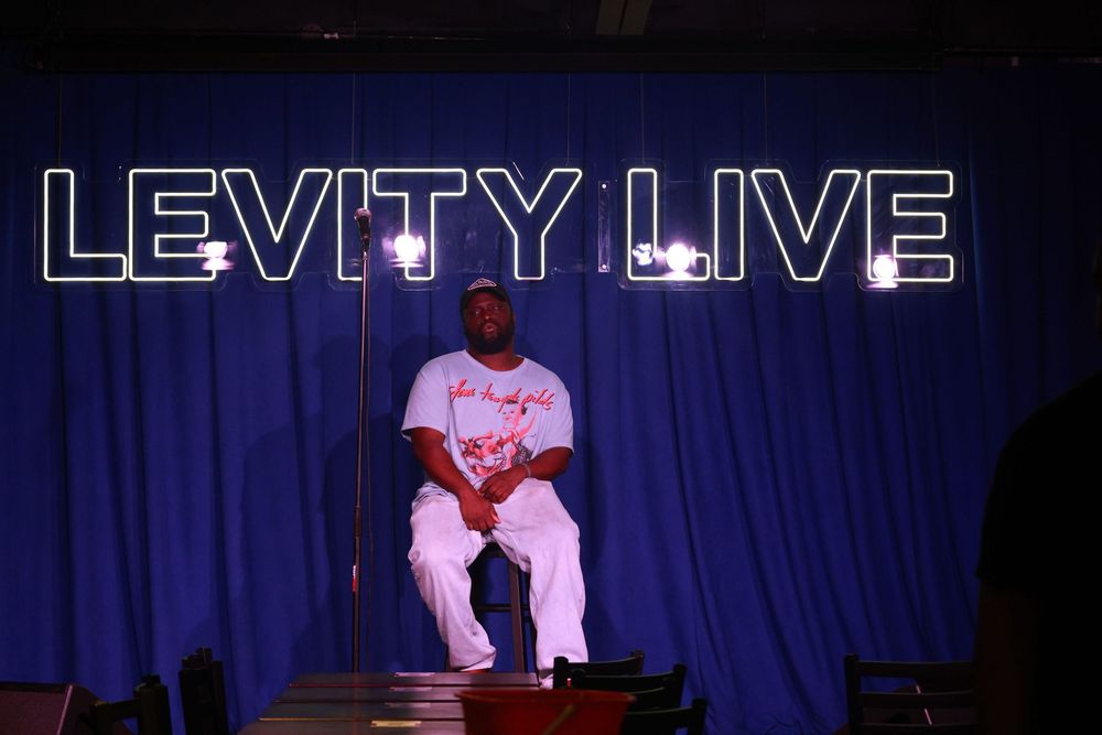 A man in white outfit sits on stage under neon Levity Live sign against blue curtain backdrop.
