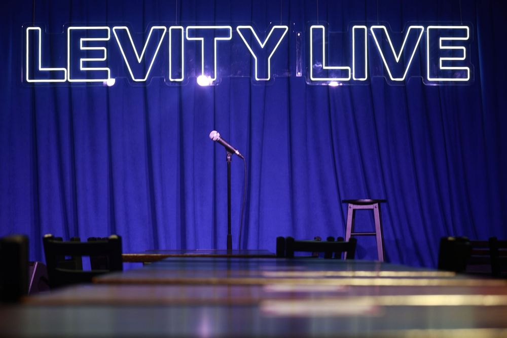 Empty Levity Live comedy club stage with microphone, stool, and blue curtain backdrop under neon sign.
