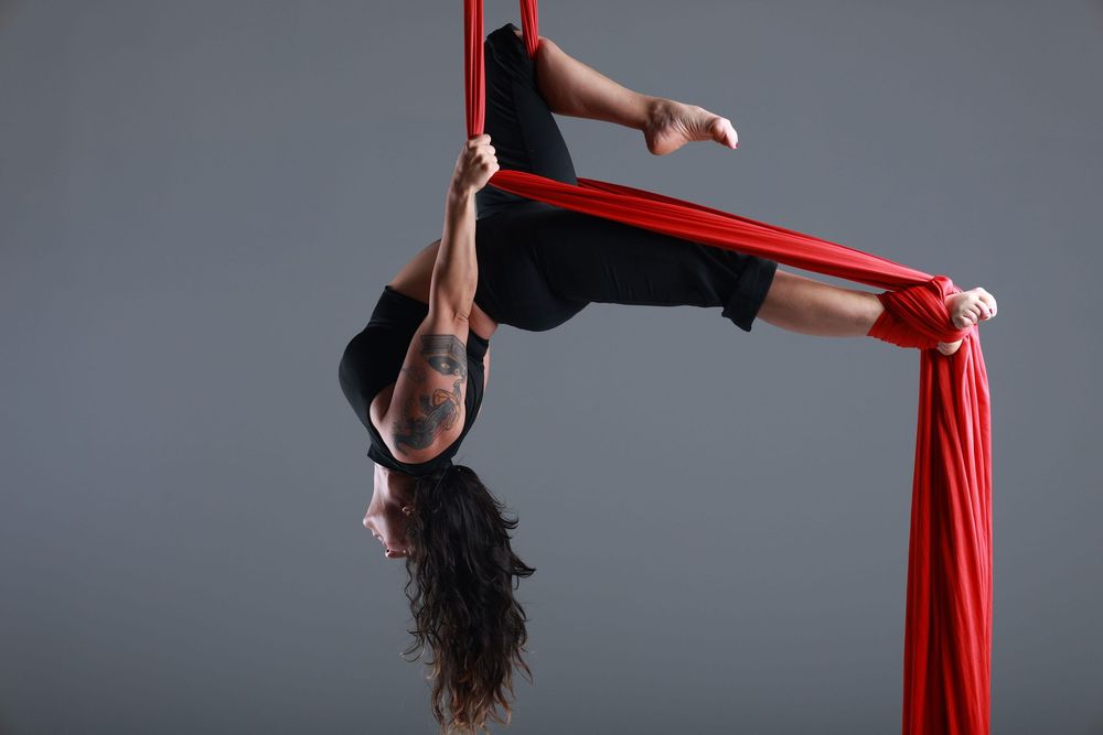 Aerial silk performer in black outfit gracefully suspended upside down on red fabric against gray studio background.