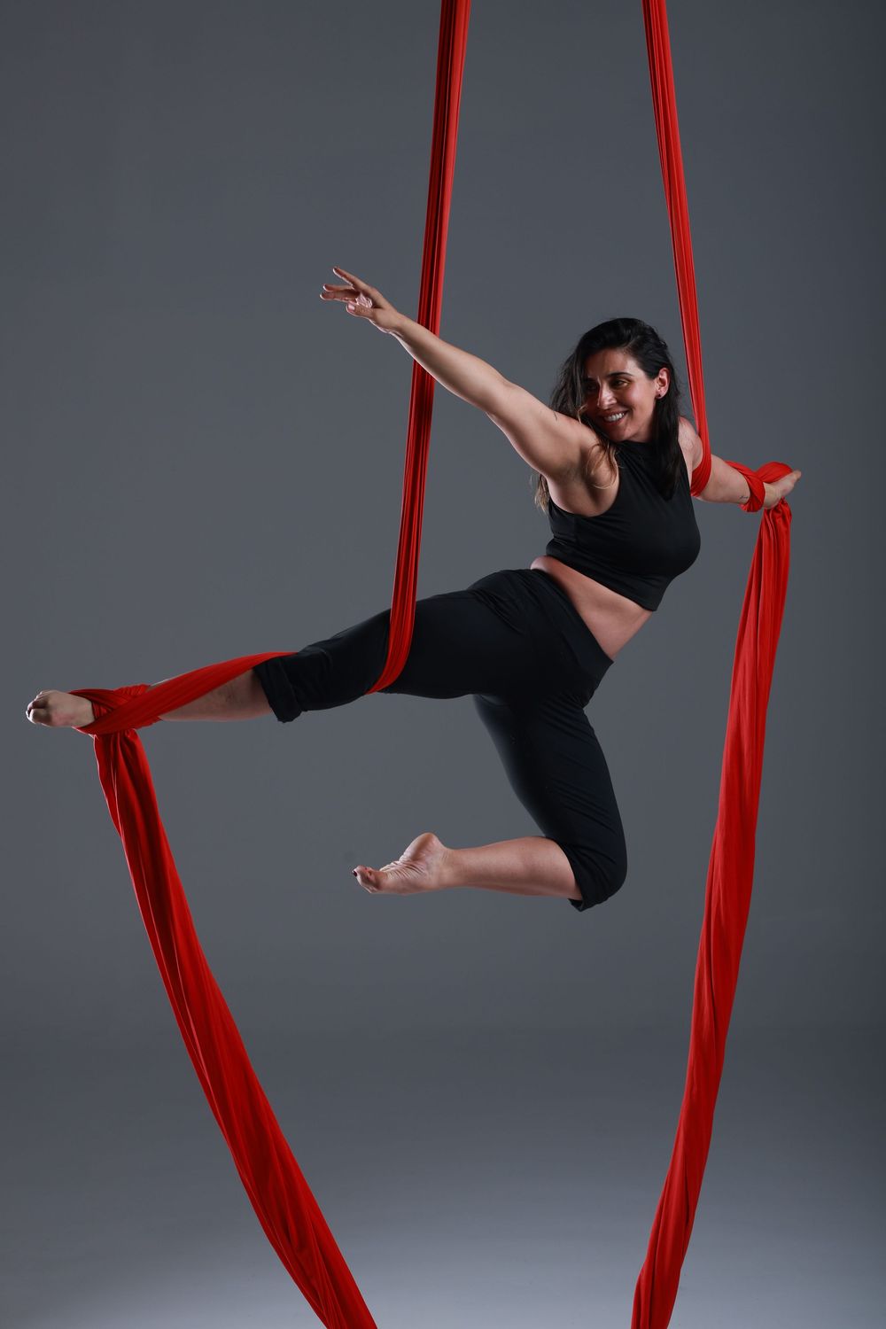 Aerial silk dancer in black outfit performing graceful pose with red silks on gray background.