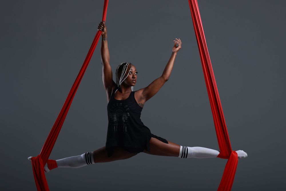 Aerial dancer performs splits on red silk ribbons wearing black outfit and white knee socks against gray background.