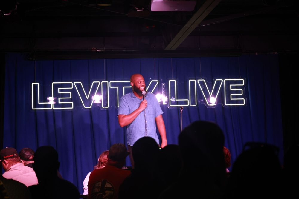 Comedian performing stand-up on stage at Levity Live comedy club under neon sign lights.