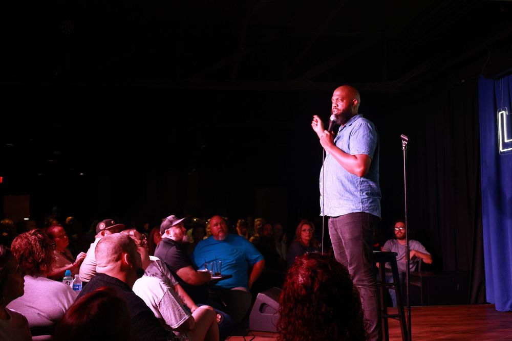 A comedian performs stand-up on stage before a large seated audience in a dimly lit comedy club.