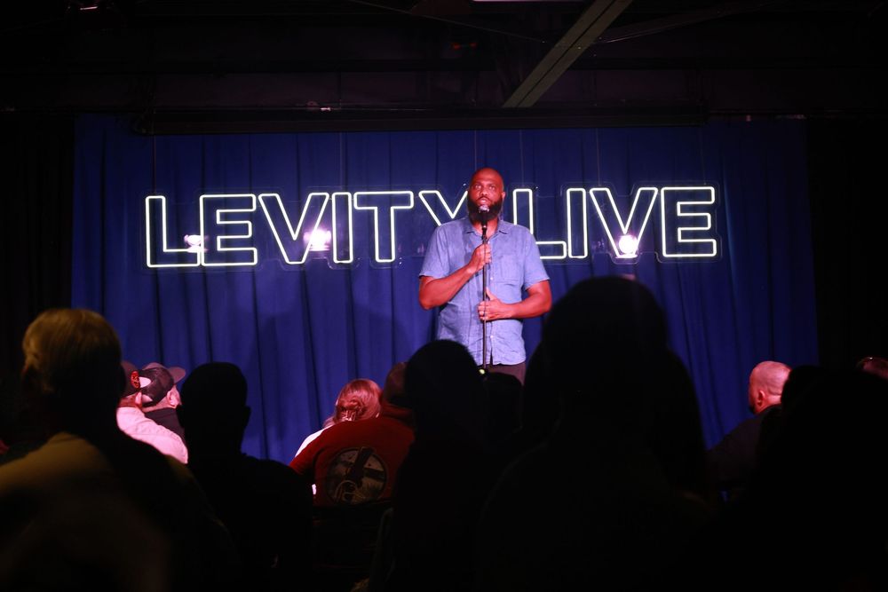 A comedian performs on stage at Levity Live comedy club, standing at a microphone before a seated audience.
