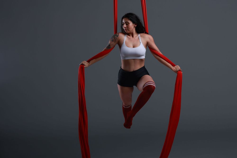 Woman performing aerial silk acrobatics, suspended in red fabric, wearing white top and black shorts on gray background.