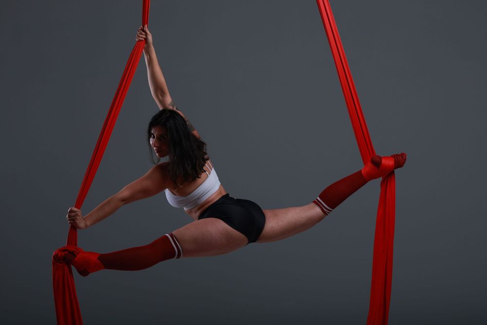 Female aerialist performing splits on red aerial silks in a studio, wearing white crop top and black shorts.