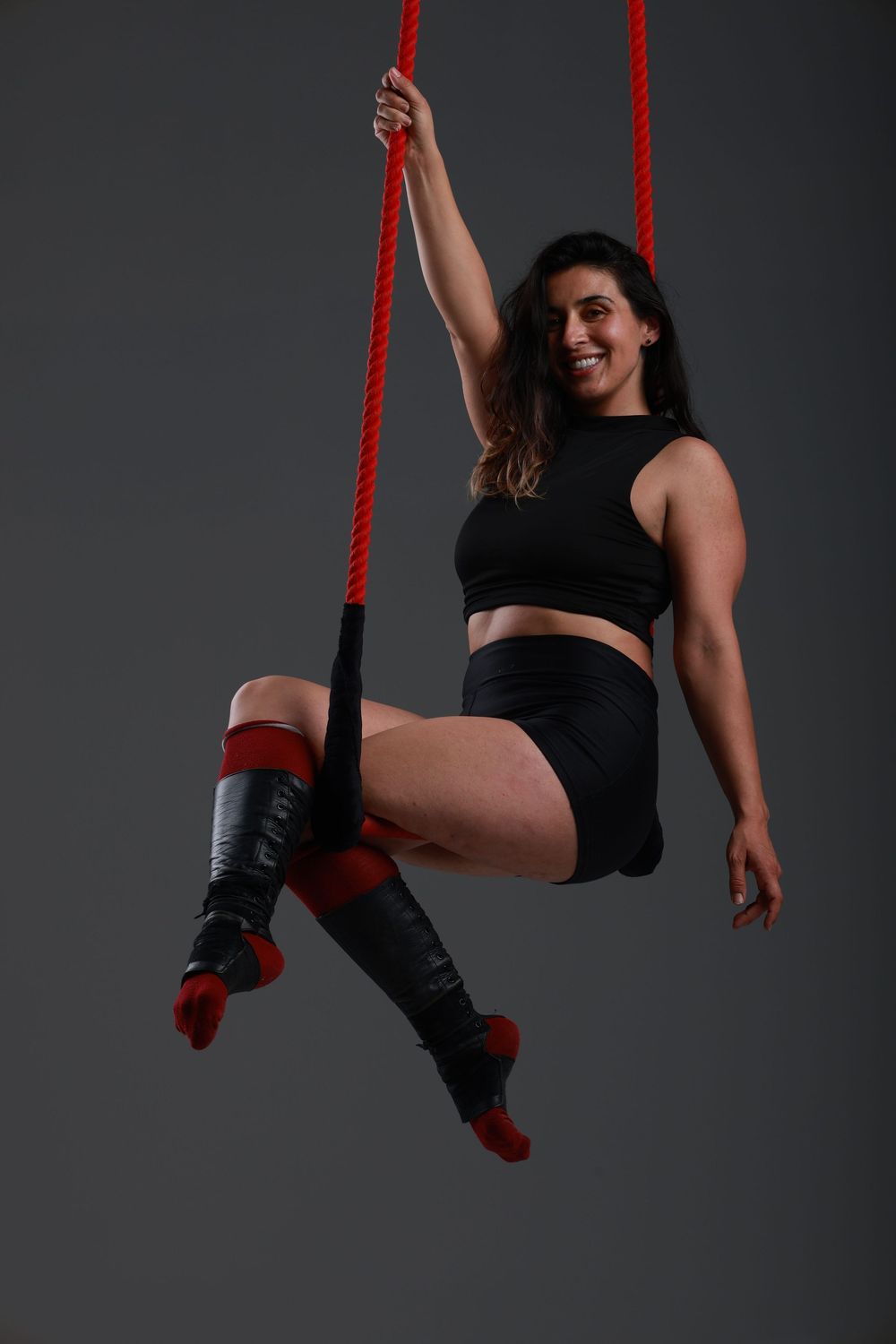 Woman in black athletic wear performs aerial acrobatics on red ropes, smiling confidently against gray background.