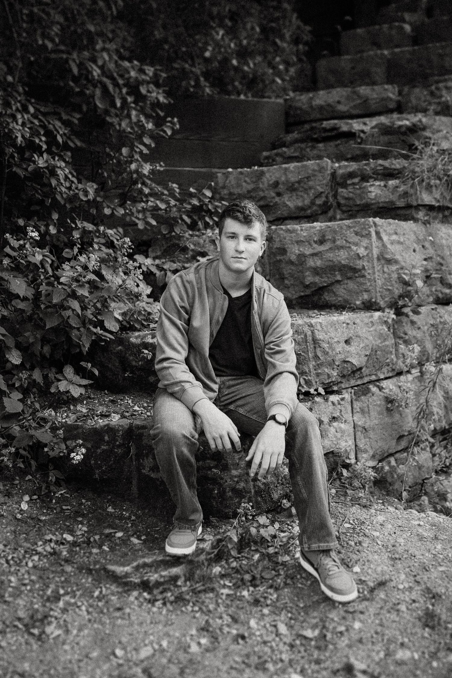 Black and white portrait of someone seated on stone steps against a rustic wall surrounded by foliage.
