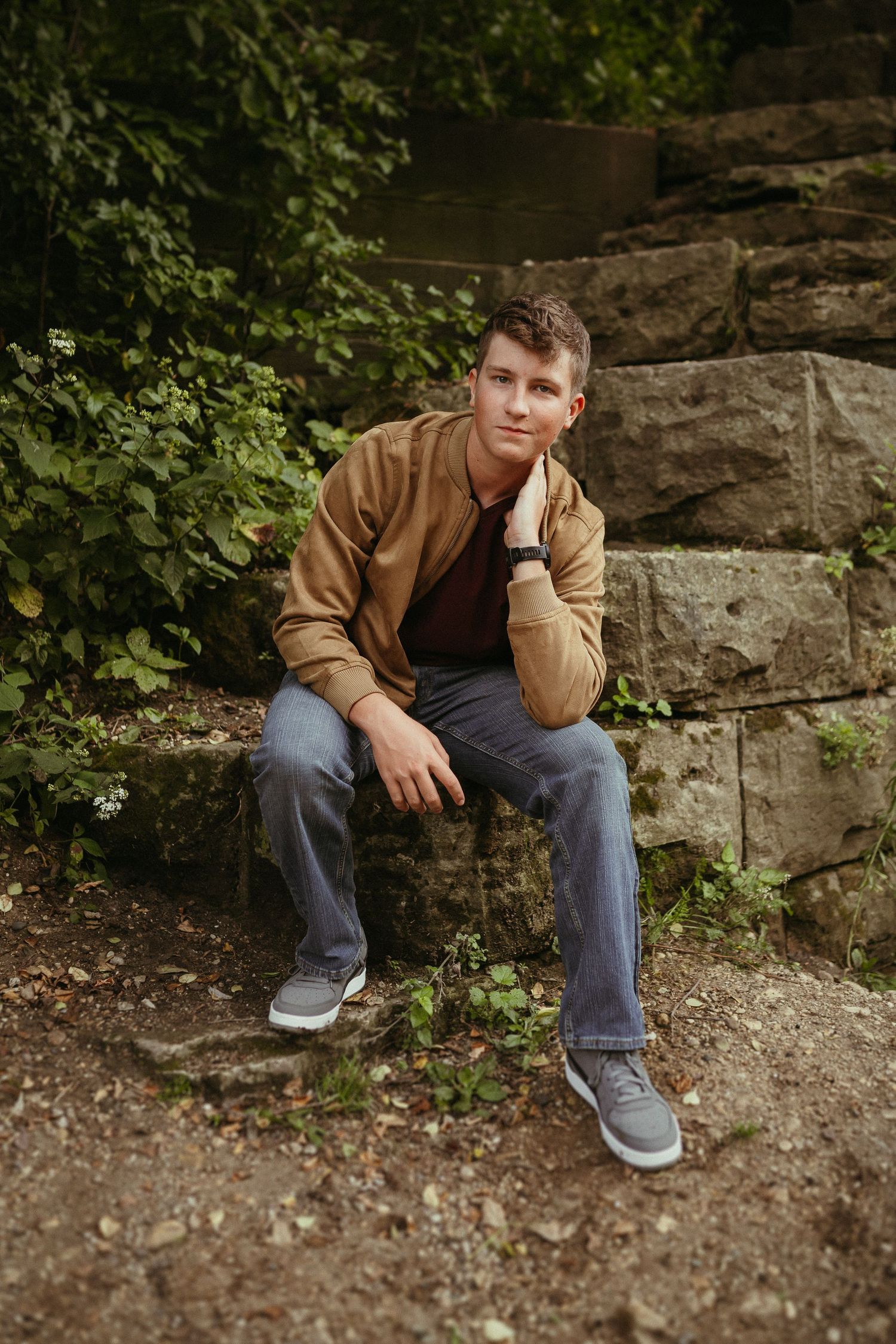 A person in a brown jacket sits casually on stone steps surrounded by greenery.
