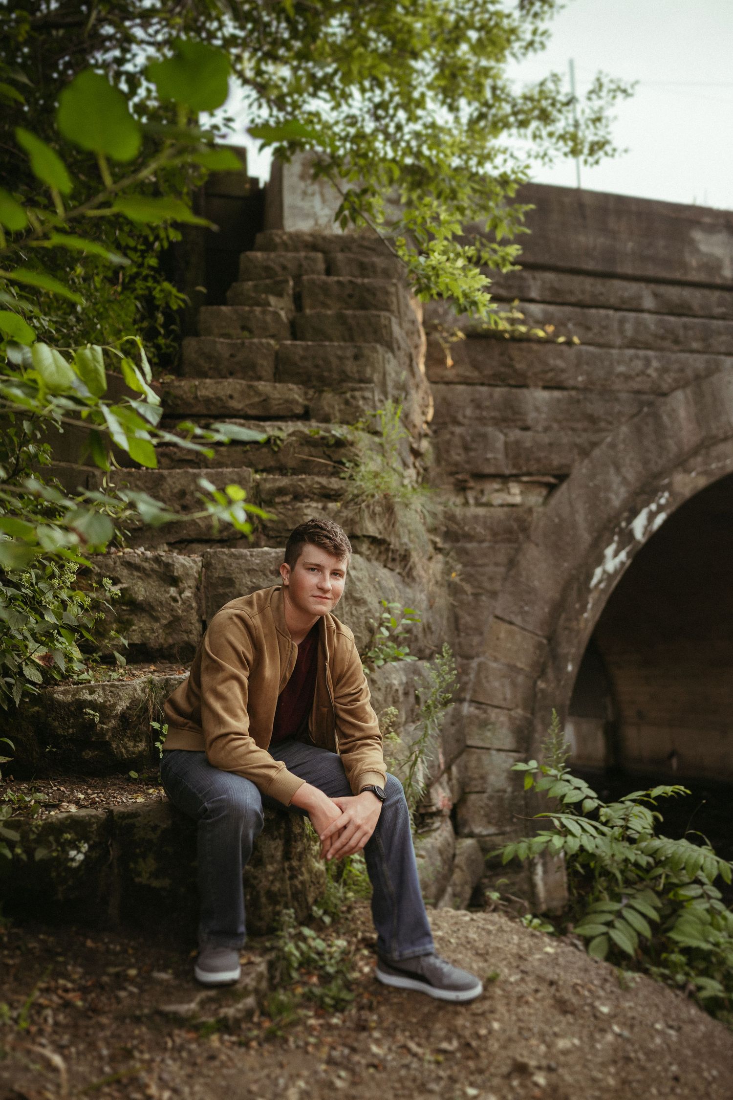 Someone sits on stone steps near an old stone bridge arch surrounded by green foliage in a tranquil outdoor setting.