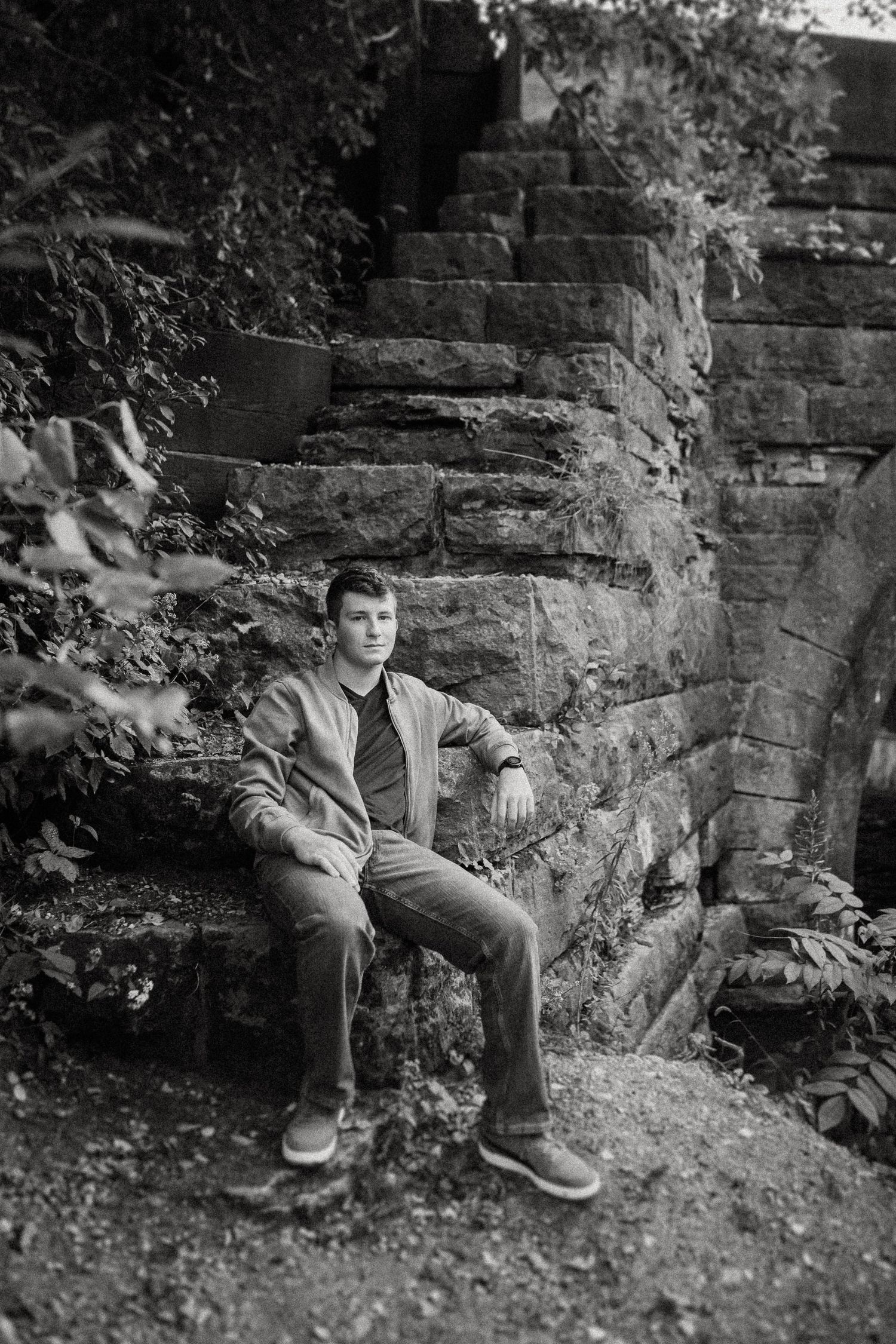 A black and white portrait of someone sitting on old stone steps against a weathered stone wall surrounded by foliage.