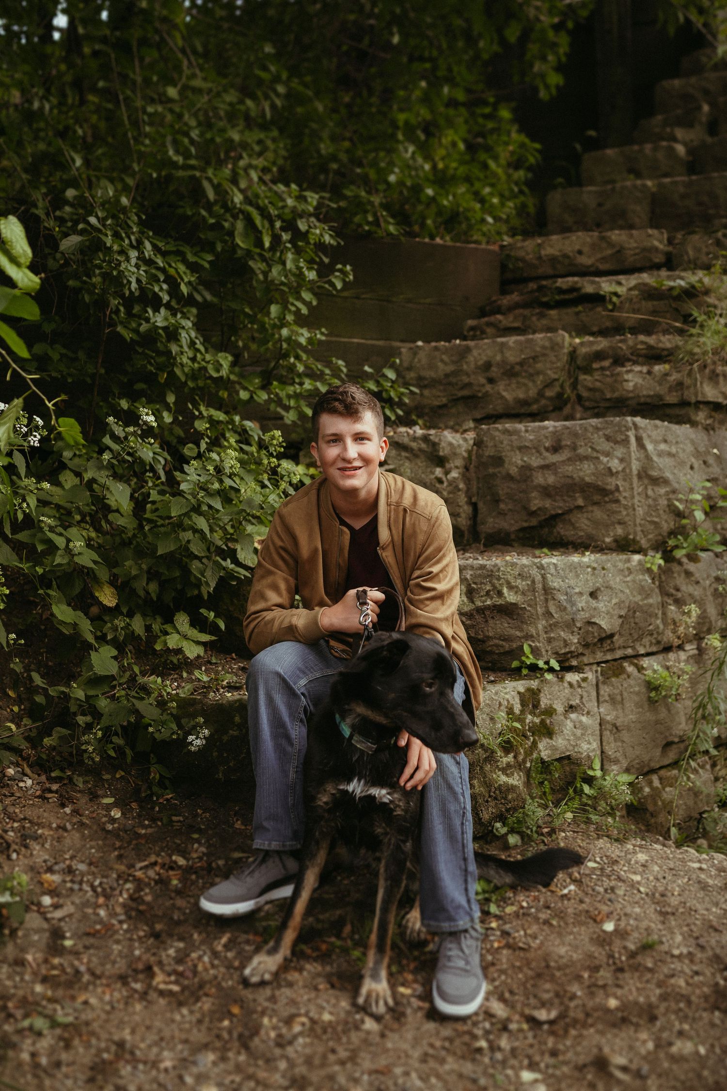 Someone in a brown jacket sits on stone steps with a black dog amongst greenery at dusk.