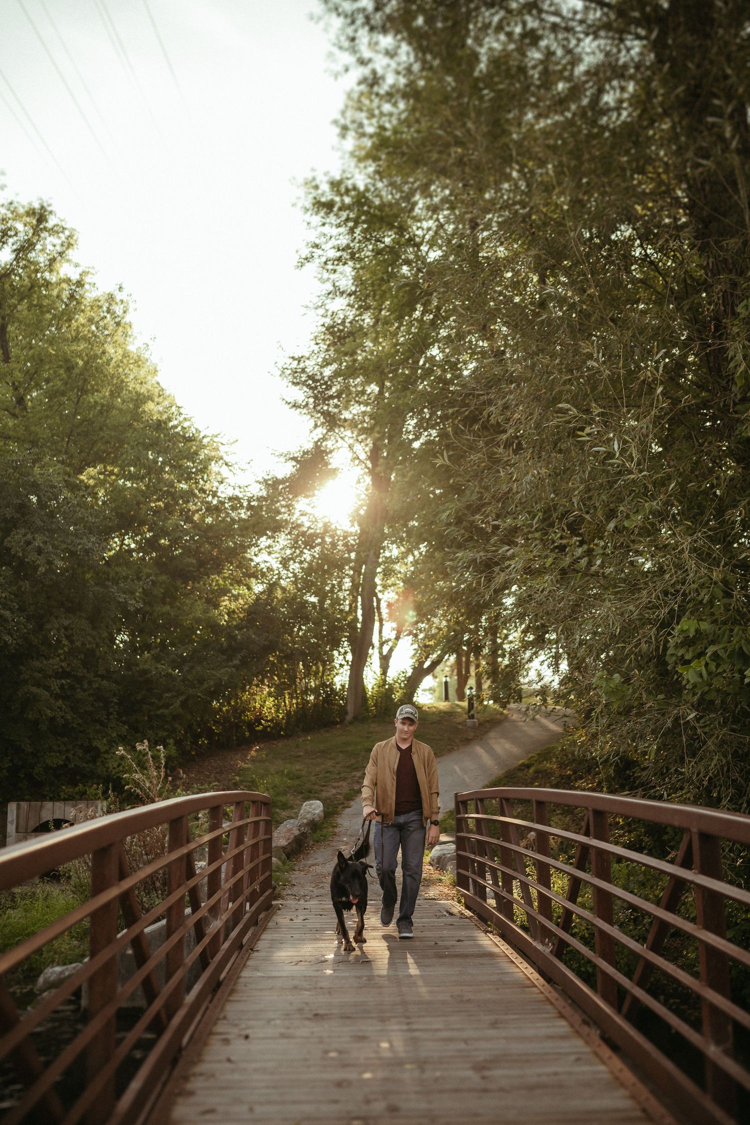 Person walking their black dog across a wooden footbridge during golden hour in a scenic wooded area.