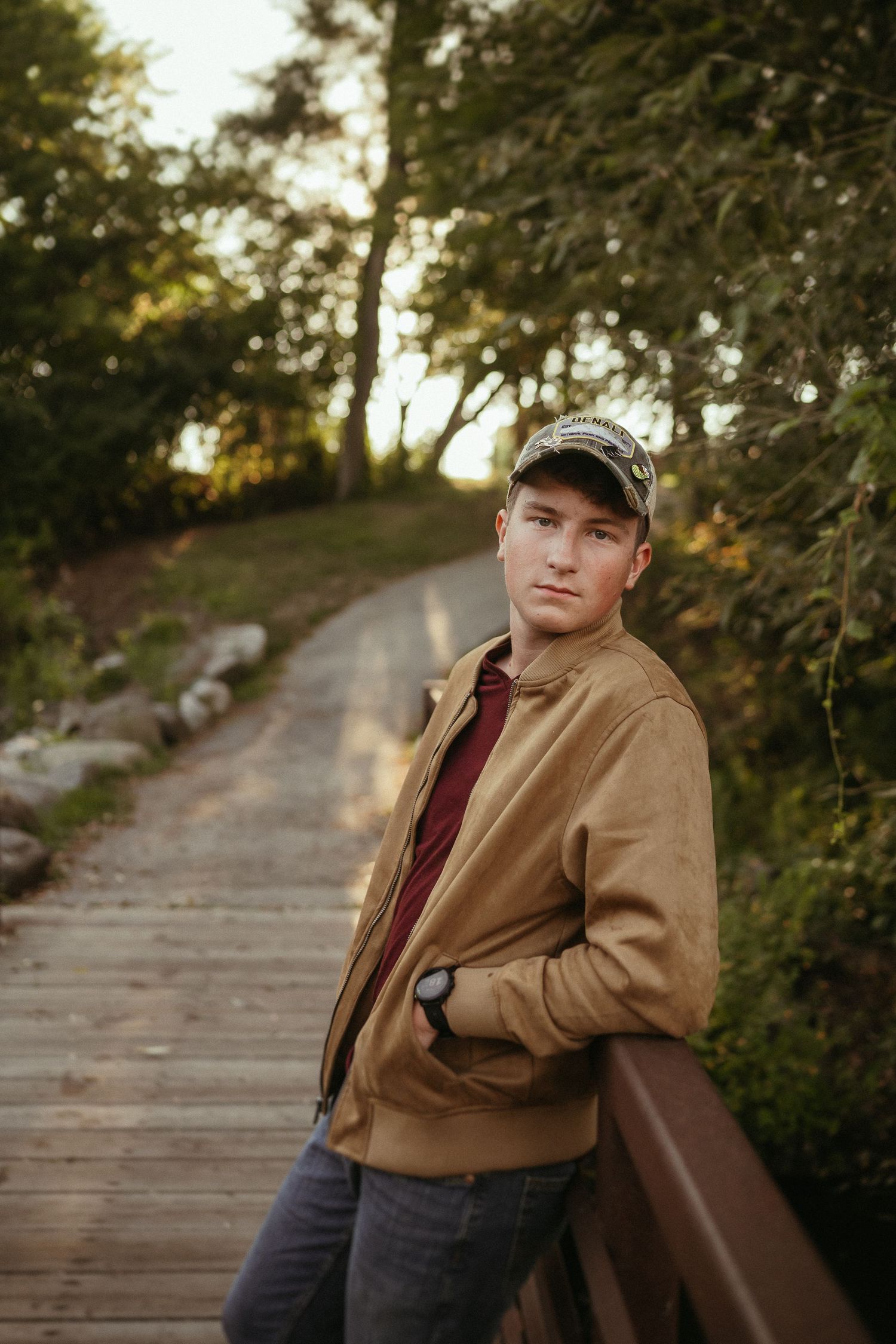 Scenic autumn boardwalk through trees with person in casual tan jacket and baseball cap during golden hour lighting.