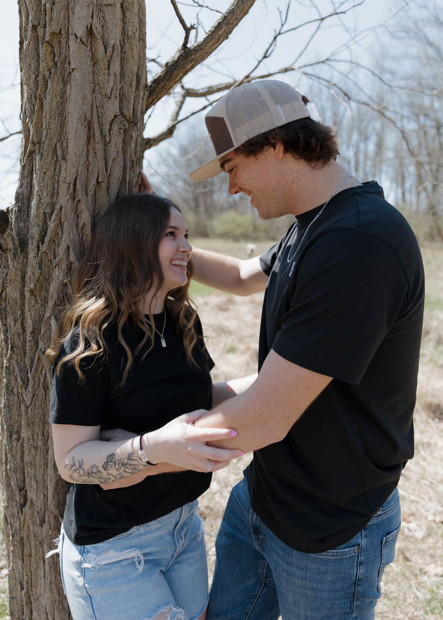 A couple in casual clothing standing close together by a tree trunk in a natural outdoor setting.