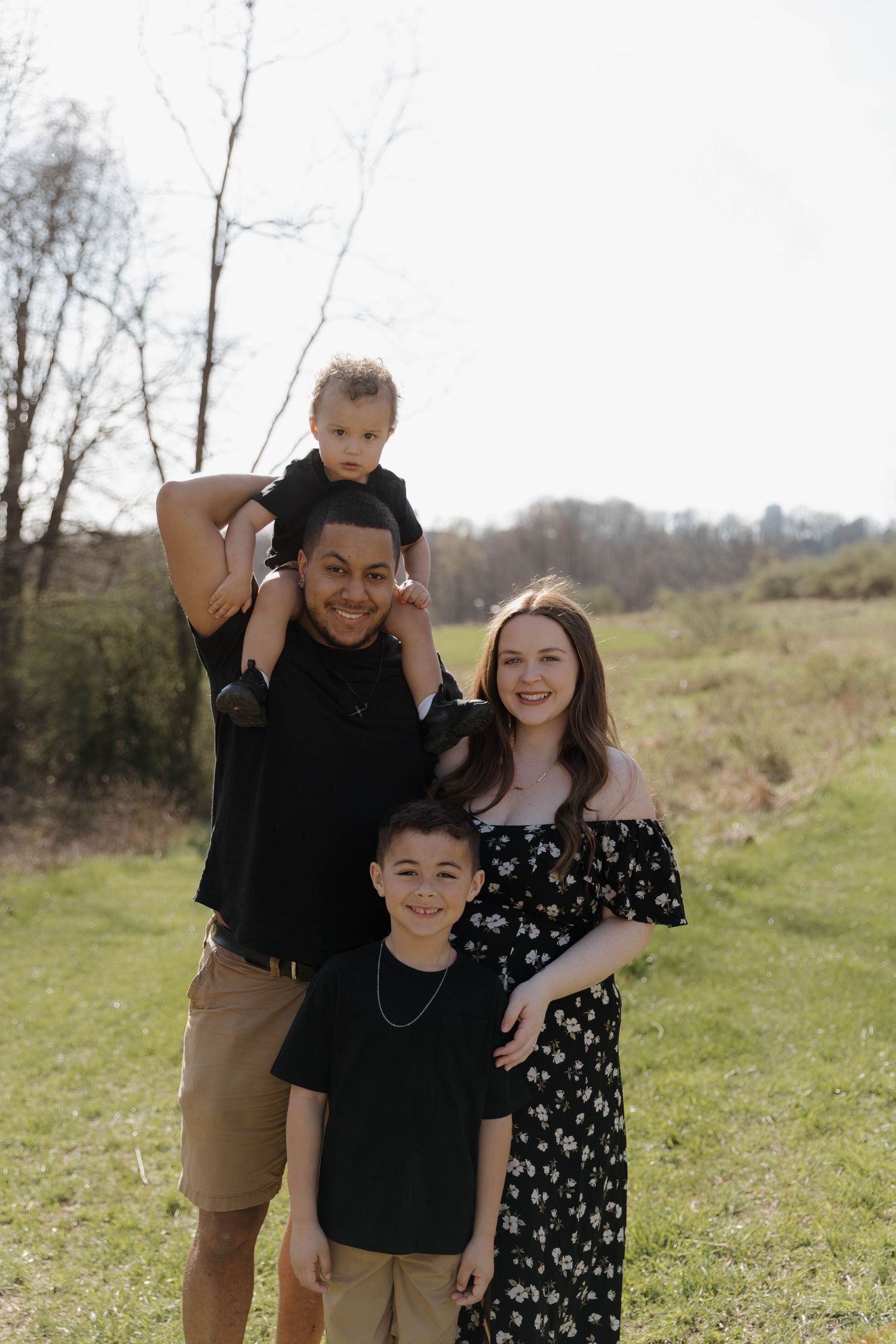 A family poses together outdoors in a grassy field with trees in the background on a sunny day.