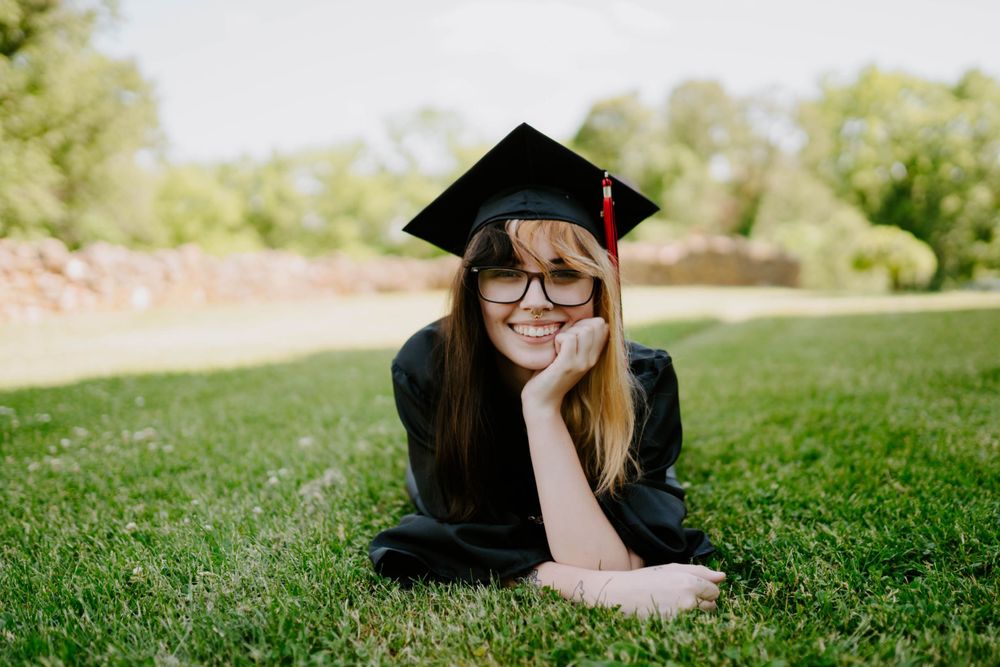 Graduate in black cap and gown lying on grass during outdoor photo session.