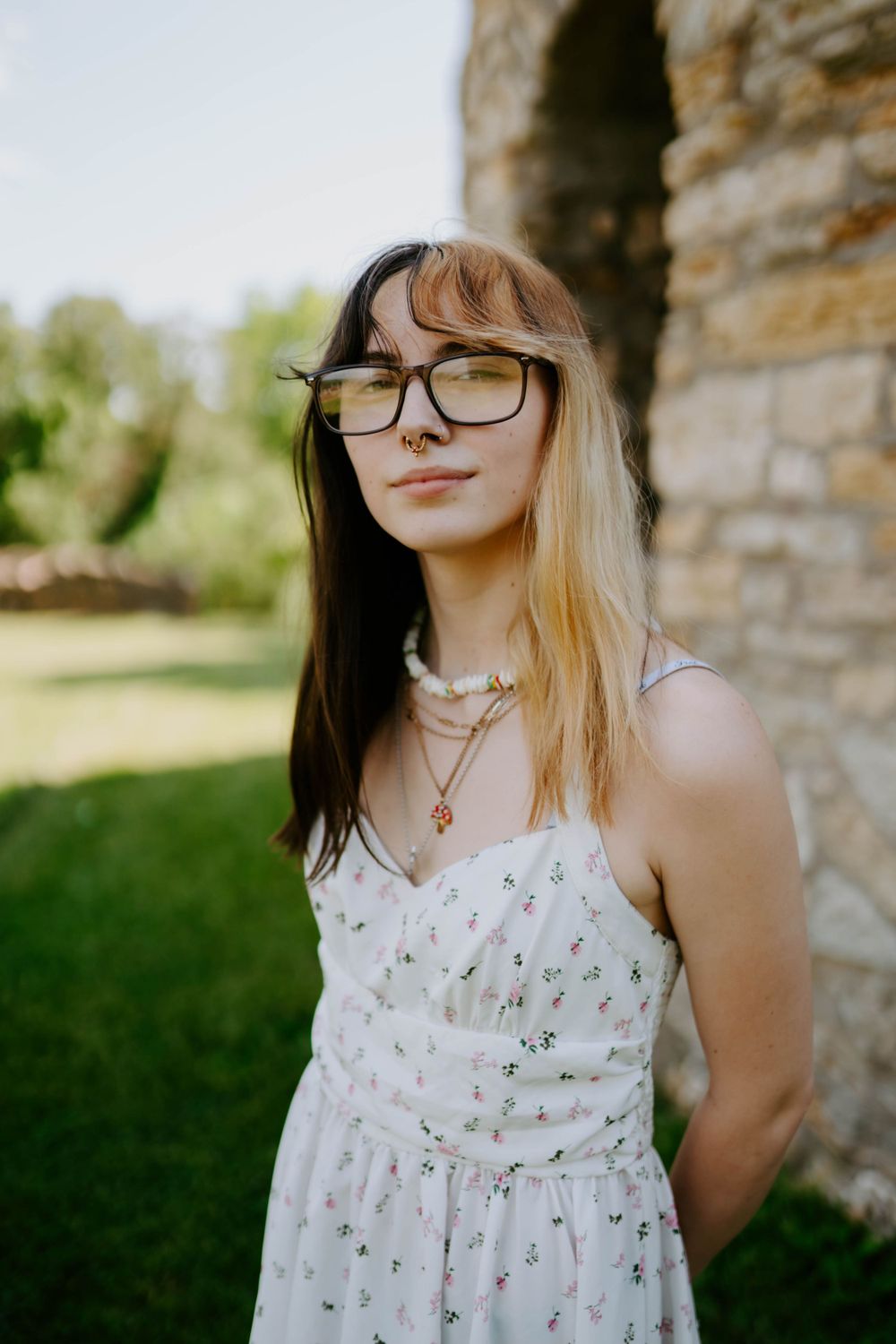 Person in white floral dress poses against stone wall in natural lighting.