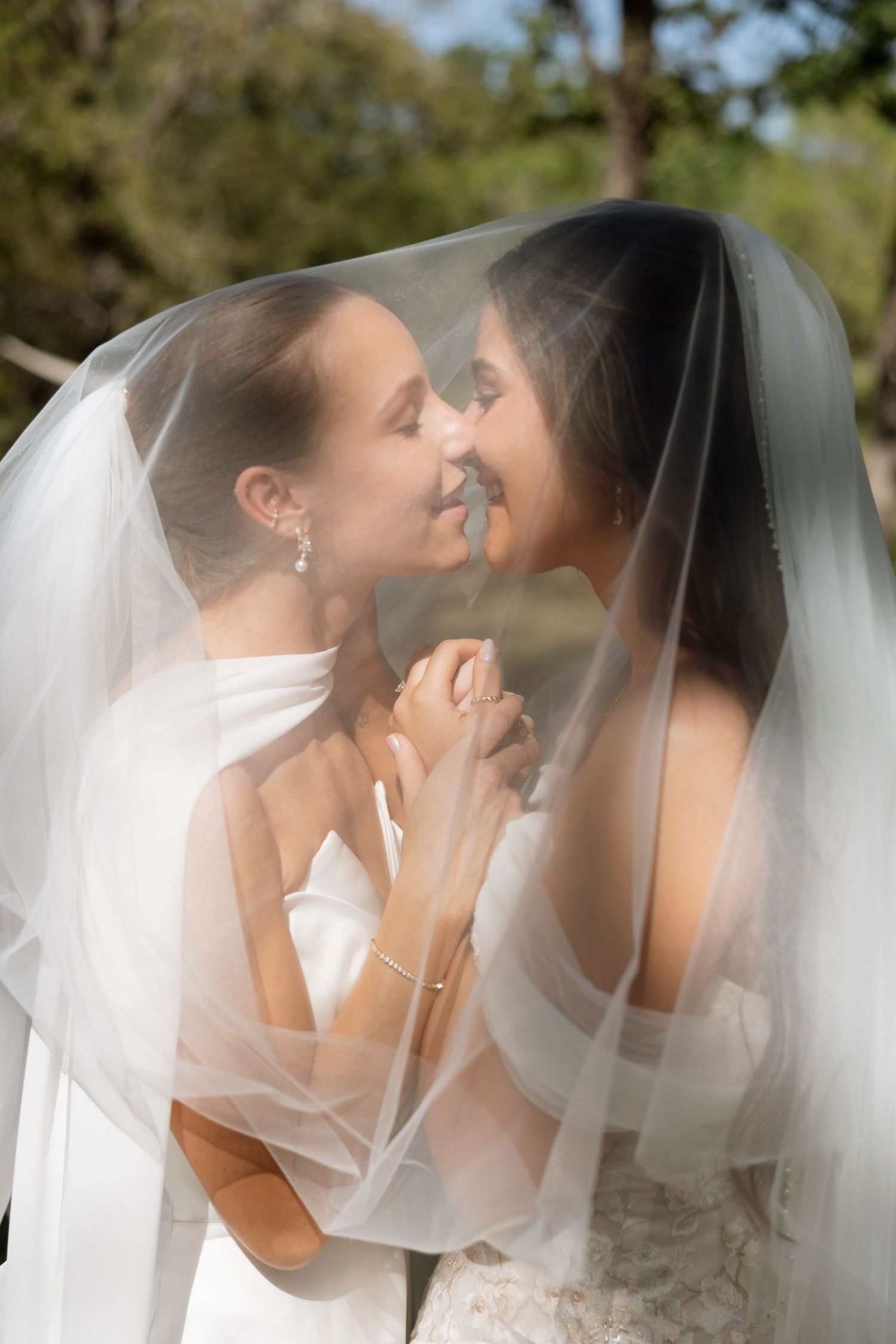 Two brides share an intimate moment together under their wedding veils in a romantic outdoor setting.