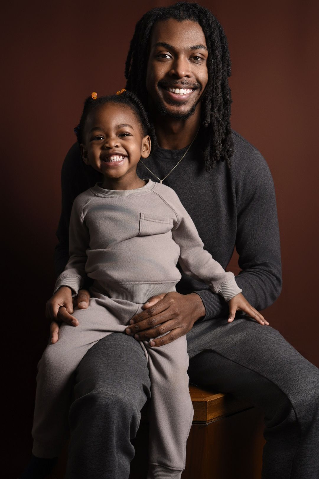 Portrait of a parent and child sitting together wearing casual grey clothing against a brown backdrop.