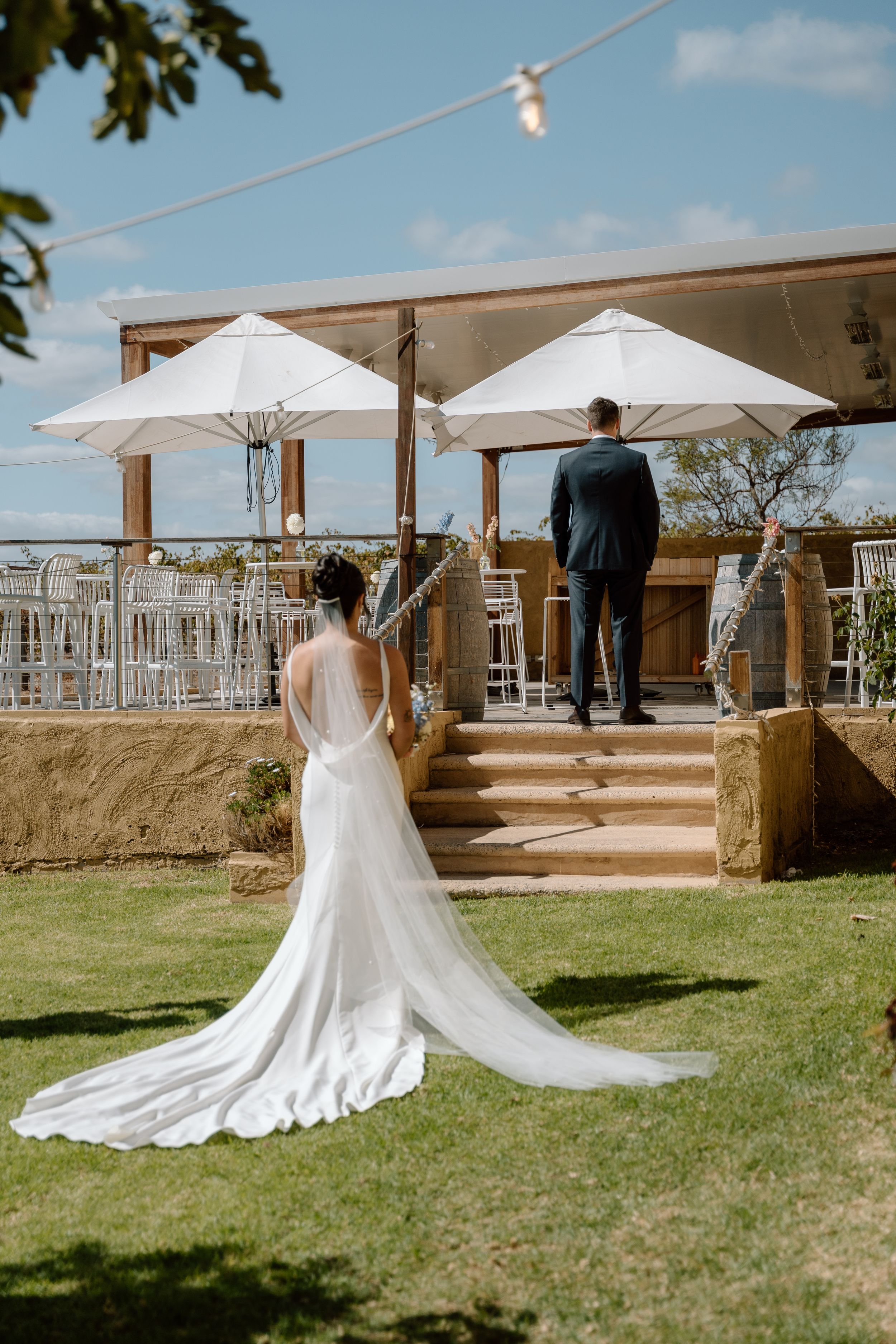 A flowing white wedding dress with a long train spreads across a grassy lawn beneath white umbrellas at an outdoor venue.