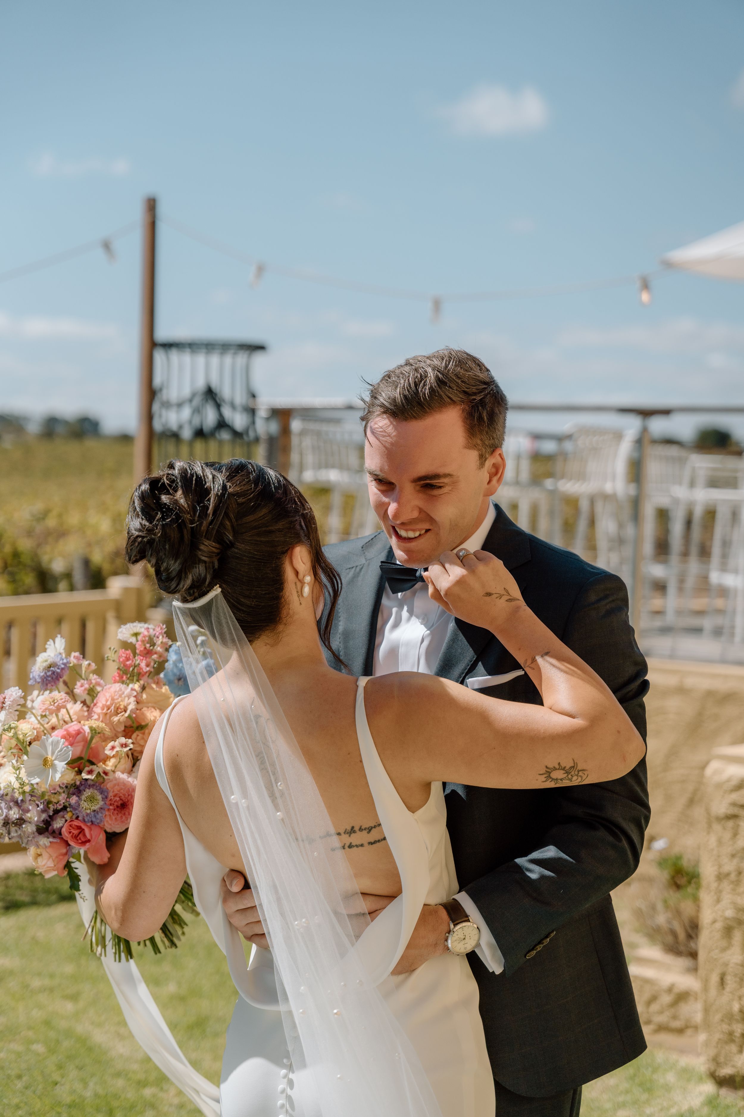 A joyful wedding couple shares a candid moment together in a sunny outdoor setting with a floral bouquet.